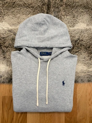 Ljusgrå Polo Ralph Lauren Fleece Hoodie - Storlek S - Hej! 👋 Säljer en riktigt skön och mjuk ljusgrå Polo Ralph Lauren fleece hoodie i storlek M. Den har den klassiska blå pony-loggan broderad på bröstet 🐎 Tröjan är i gott skick och känns väldigt fräsch. Jag vill dock vara helt transparent med att det finns två mycket små hål/slitningar på ryggen (se närbild). Det är inget som man tänker på vid användning, men priset är satt därefter! ✨ 100% äkta med både tvättlappar och QR-kod kvar för enkel verifiering ✅ Ett perfekt och bekvämt premium-basplagg. 