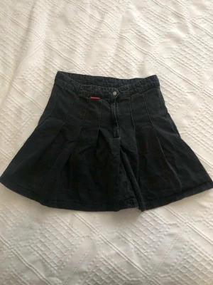 Svart kjol vintage - Cool svart skort  med breda ben och markerade sömmar. Gjord i denim med knapp och dragkedja framtill. Perfekt för dig som vill ha kjol-looken men ändå känna dig bekväm. Röd logotypetikett i midjan.