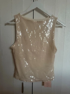 Beige paljettlinne - Snyggt beige linne täckt med glittrande paljetter. Ärmlös modell med rund halsringning och croppad passform. Perfekt för dig som vill sticka ut lite extra.
