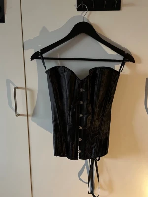 Svart satin korsett med snörning - Snygg svart korsett i glansigt satinmaterial. Framsidan har metallknäppning och baksidan snörning för justerbar passform.
