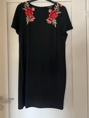 Svart klänning med blommor från Lipsy - Svart kortklänning från Lipsy med broderade röda och rosa blommor vid axlarna. Klänningen har korta ärmar och en rak, enkel passform. Perfekt för dig som gillar stilrena plagg med en färgstark detalj.