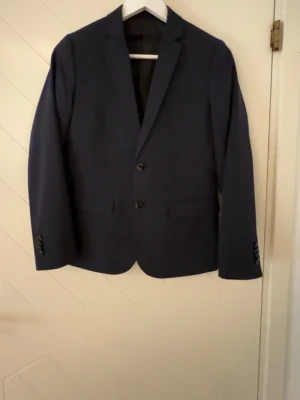 Mörkblå kavaj med två knappar - Säljer en stilren mörkblå kavaj med klassisk passform och två knappar framtill. Kavajen har två fickor med lock och knappar på ärmsluten. Perfekt för dig som vill ha en enkel och snygg look. Materialet känns mjukt och bekvämt.