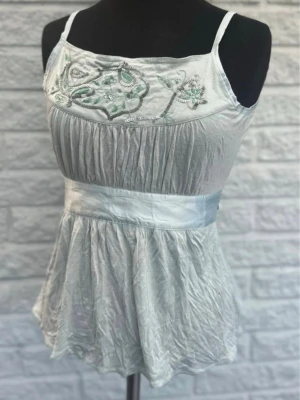 Ljusturkos broderad linnetopp - Supersöt ljusturkos linnetopp med tunna axelband och broderade detaljer i grönt och silver framtill. Toppen har en markerad midja med bred satinliknande kant och ett lätt glansigt, skirt tyg som ger en flowy känsla.