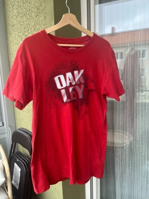 Röd Oakley t-shirt med tryck - Röd t-shirt från Oakley med stort vitt och svart tryck på bröstet. Modellen är regular fit och har rund hals samt korta ärmar. Perfekt för en avslappnad och sportig stil.