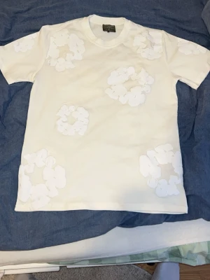 Vit t-shirt med blommor från Denim Tears storlek L - Säljer en vit t-shirt från Denim Tears med unika vita blommönster tryckta över hela tröjan. T-shirten har rund hals och klassisk passform. Perfekt för dig som gillar streetwear och vill sticka ut med en stilren men ändå speciell look.