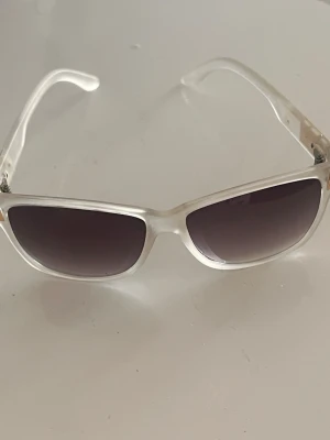 Vita kantiga solglasögon - Snygga solglasögon med vit, transparent båge och mörka glas. Modellen har en kantig form som ger en cool och trendig vibe. Perfekta för att lyfta din stil under soliga dagar.