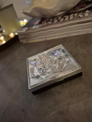 Lancome highlighter ros - Exklusiv highlighter från Lancome i en lyxig ask med kristalliknande lock och rosformad design i pudret. Färgen är skimrande vit med pärlemor-effekt som ger en snygg glow till huden. Perfekt för att framhäva kindben och ge lyster till ansiktet. Kan vara Testad/swatchad. Lyxig förpackning