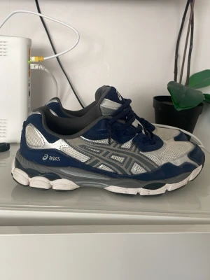 Asics blå och silver sneakers - Snygga sneakers från Asics i blått och silver med mesh- och syntetdetaljer. Skorna har en sportig vibe med klassisk Asics-logga på sidan och tungan. Perfekta för dig som gillar streetwear och bekväma skor med cool design.