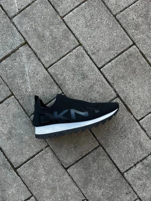 Svarta slip-on sneakers från DKNY - Svarta sneakers från DKNY med slip-on design och tydlig logga på sidan. Ovandelen är i textil och sulan är vit med svarta detaljer och sågtandsmönster undertill. Snygg och modern siluett som passar till streetwear.