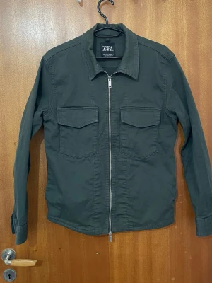 Mörkgrön overshirt från Zara - Stilren mörkgrön overshirt från Zara med raka linjer och två stora bröstfickor. Dragkedja framtill och klassisk krage ger en clean look. Tillverkad i ett mjukt bomullstyg som känns bekvämt och passar perfekt till lager på lager-stil.