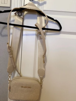 Beige axelremsväska från Valentino - Stilren beige handväska från Valentino med bred justerbar axelrem och präglad logotyp på framsidan. Väskan är rektangulär och tillverkad i ett strukturerat konstlädermaterial. Guldiga metalldetaljer och dragkedja upptill ger en lyxig känsla. (endast använd en gång, har inte vågat använda den mer pga kostnaden av en märkesväska. är äkta och köpt för ett år sedan)
