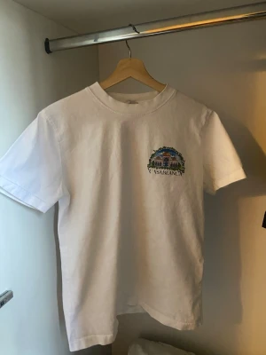 Vit Casablanca t-shirt med tryck - Vit t-shirt från Casablanca med färgglatt tryck av byggnader och texten 'CASABLANCA' på bröstet samt stort motiv och texten 'CASA BLANCA' på ryggen. Klassisk passform och rund hals. Perfekt för dig som gillar unika prints och streetwear.