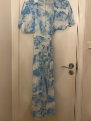 Blåvit långklänning från Gina Tricot - Långklänning från Gina Tricot i blått och vitt med omlottmodell och korta, vida ärmar. Klänningen har ett unikt mönster som påminner om moln och är tillverkad i ett lätt och luftigt material, perfekt för varma dagar.