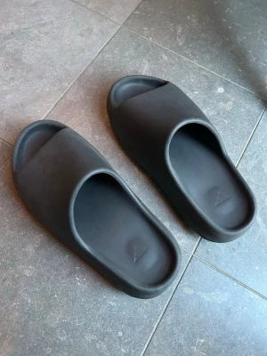 Svarta Yeezy Slide tofflor - Säljer ett par svarta Yeezy Slide tofflor från Adidas. De har en chunky design med bred rem över foten och räfflad sula för extra grepp. Tillverkade i mjukt och flexibelt skumgummimaterial som gör dem riktigt bekväma att glida runt i.