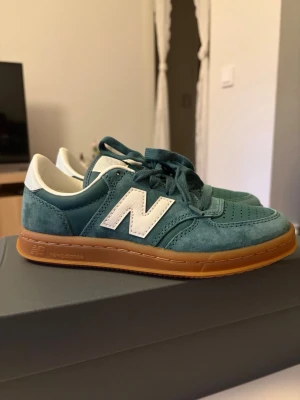 New Balance teal sneakers med gummisula - Snygga tealblå New Balance sneakers i mocka och textil med vit logga på sidan och vit häl. Skorna har en klassisk låg profil och brun gummisula som ger en retrovibe. Perfekta för dig som gillar streetstyle och vill sticka ut med färg.