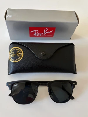 Svarta Ray-Ban Clubmaster solglasögon - Snygga svarta Ray-Ban Clubmaster solglasögon med klassisk halvbåge och mörka glas. Kommer med original svart fodral och box. Perfekt accessoar för att lyfta din stil och skydda ögonen mot solen.