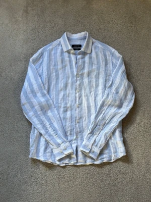 Randig linneskjorta från Massimo Dutti - Snygg långärmad skjorta i ljusblått och vitt från Massimo Dutti. Skjortan är tillverkad i 100% linne och har klassisk krage, knäppning framtill och randigt mönster. Perfekt för en avslappnad och stilren look.