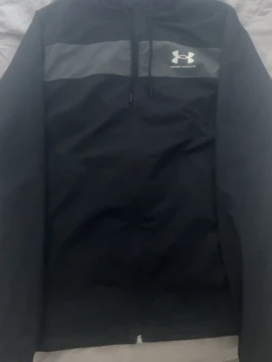 Under Armour vindjacka  - Svart vindjacka från Under Armour med grått bröstparti och logga på vänster sida. Jackan har huva med dragsko och dragkedja framtill. Tillverkad i lätt och slitstarkt material, perfekt för blåsiga dagar. Storlek S, helt nytt och pris kan diskuteras 