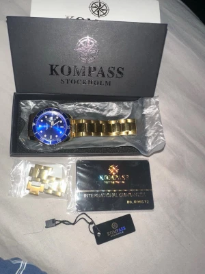 Kompass Stockholm guld klocka - Snygg klocka från Kompass Stockholm med guldigt armband i metall och blå urtavla. Kommer i originalask med extra länkar och garanti. Klockan har en klassisk rund form och stilren design som sticker ut. Aldrig använd bara bytt storlek till mindre. Skriv om funderingar eller om att diskutera pris