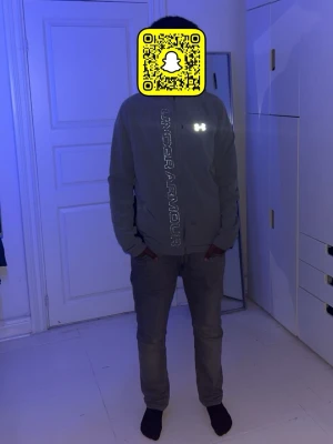 Grå vindjacka från Under Armour - Snygg grå vindjacka från Under Armour med reflekterande logga på bröstet och stort tryck längs dragkedjan. Jackan har huva, hel dragkedja och svarta detaljer på insidan. Perfekt för blåsiga dagar och sportiga outfits.