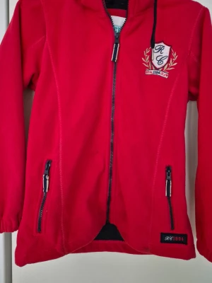 Fleecejacka - Röd fleecejacka från mywear Active med huva, hel dragkedja framtill och två fickor med dragkedjor. Jackan har broderad logga på bröstet och elastiska ärmslut. Perfekt för kyliga dagar. Den har några fläckar.
