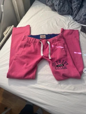 Rosa mjukisbyxor från Superdry - Snygga rosa mjukisbyxor från Superdry med vitt dragsnöre i midjan och broderad text 'Superdry J.P.N College' på ena benet. Byxorna har två sidofickor och är gjorda i mjukt bomullsmaterial. Midjemått rakt av: 40cm Innerbenslängden: 78cm