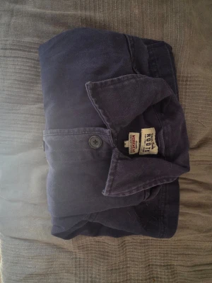Mörkblå overshirt från Roots - Säljer en stilren mörkblå overshirt från Roots med klassisk krage och knäppning framtill. Overshirten har två stora fickor framtill och är tillverkad i ett mjukt bomullstyg. Perfekt att slänga över en t-shirt för en avslappnad look.