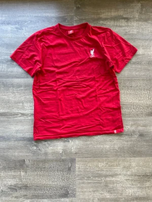 Röd Liverpool FC t-shirt - Röd t-shirt från Liverpool FC med klubbens logga broderad i vitt på bröstet. Tröjan är 100% bomull. Passar dig som är Liverpool supporter! Skriv vid frågor