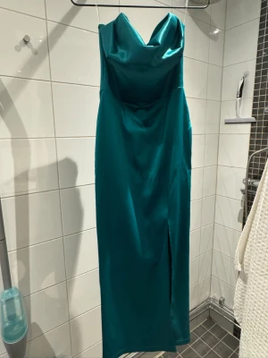 Turkos långklänning i satin - Snygg turkos långklänning i satin med draperad urringning och slits framtill. Klänningen är ärmlös och har en elegant, figurnära passform. Perfekt för dig som vill sticka ut med en stilren och modern look.