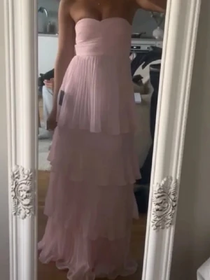 Ljusrosa axelbandslös balklänning - helt ny ljusrosa balklänning med volanger i flera lager och axelbandslös bandeau-topp. Klänningen har en flowy passform och är tillverkad i ett tunt, plisserat material som ger en drömmig känsla. passade ej, nypris 1300