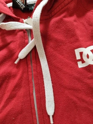 Röd DC zip-hoodie med vita detaljer - Säljer en röd hoodie från DC Shoes med dragkedja, vit logga på bröstet och vita snören i huvan. Ärmsluten har vita och röda ränder för en sportig look. Perfekt för dig som gillar streetwear och skate. Materialet är mjuk bomull.