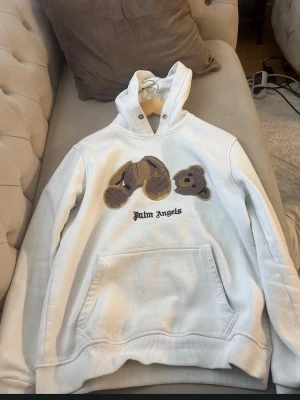 Vit hoodie från Palm Angels med björn - Säljer en vit hoodie från Palm Angels med en ikonisk björn-applikation på bröstet och svart text. Hoodien har huva, magficka och ribbade muddar. Perfekt för dig som gillar streetwear och vill sticka ut med en unik design.