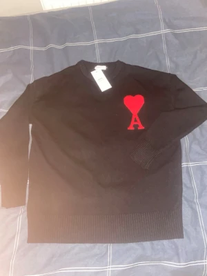  Ami Paris - Svart stickad tröja från Ami Paris med rund halsringning och ribbade muddar. På bröstet finns ett rött hjärta med bokstaven A, vilket är varumärkets ikoniska detalj. Perfekt för dig som gillar stilrena och trendiga plagg.