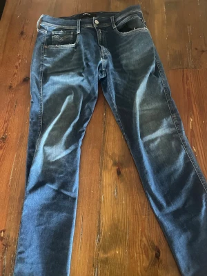 Blå jeans från REPLAY  - Säljer ett par klassiska blå jeans från REPLAY med fem fickor och snygga slitningar framtill. Jeansen har raka ben och tydliga sömmar på bakfickorna. Perfekta för dig som gillar en tidlös denimlook.