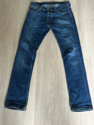 Nudie grim tim - Snygga mörkblå slim fit jeans från Nudie med klassiska orange sömmar och femficksdesign. Jeansen har en lätt tvättad look och raka ben. Perfekta för dig som gillar en stilren och modern denimstil. 32/32