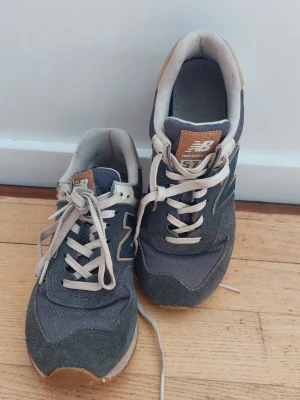 New Balance 574 mörkblå sneakers - Klassiska New Balance 574 sneakers i mörkblå mocka och textil med beige detaljer och vit sula. Skorna har den ikoniska N-loggan på sidan och bruna detaljer på plösen och hälen. Perfekta för en avslappnad och sportig stil.