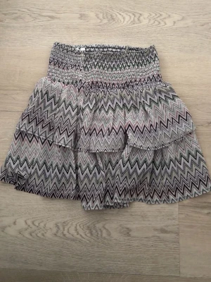 Mönstrad kortkjol med volanger - Säljer en kort kjol med volanger. Kjolen har ett snyggt zickzack-mönster i svart, vit och lila, grönt. Perfekt för dig som gillar färg och mönster. Lätt och luftig modell som passar till många olika stilar.