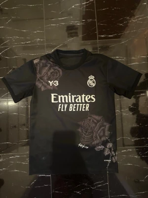 Real Madrid x Y-3 svart matchtröja - Snygg svart matchtröja från Real Madrid i samarbete med Y-3. Tröjan har vita tryck med klubbmärke, sponsorlogga och Y-3-logga samt unika rosdetaljer på axel och nederkant. Tillverkad i lätt och funktionellt material, perfekt för fotboll eller träning.