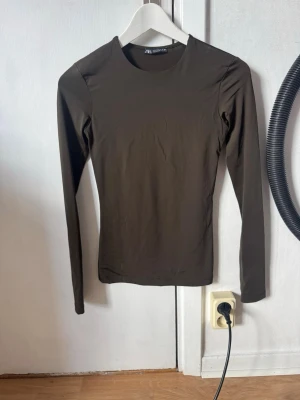 Mörkkhaki långärmad topp från Zara - En stilren khaki långärmad topp från Zara med rund halsringning. Toppen har en slim passform och är tillverkad i ett stretchigt syntetmaterial som sitter snyggt på kroppen. Perfekt att styla med jeans eller kjol.