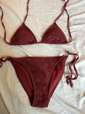 Vinröd bikini från Waikani med knyt - Säljer en vinröd bikini från Waikani i storlek XS. Bikinin har virkade detaljer och knyt i sidorna på underdelen samt i nacken och ryggen på överdelen. Perfekt för sommaren och strandhäng!,använt typ en gång 