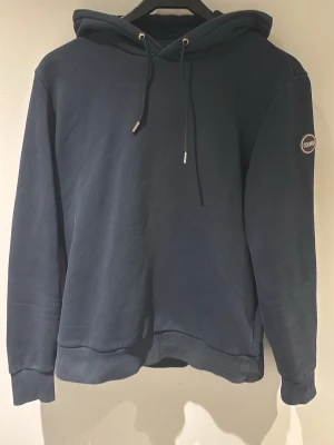 Mörkblå colmar hoodie med snörning - Mörkblå colmar hoodie med huva och snörning, ribbade muddar och ett diskret märke på ärmen. Klassisk och enkel design som passar till det mesta. Tillverkad i mjuk bomull för skön känsla. Storleken ligger mellan small och medium. På mig som varierar mellan medium och large är tröjan lite liten på. Inga defekter men lite ”tvättad” look. Hör av dig vid minsta fundering!