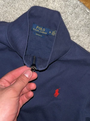 Mörkblå zip-tröja från Polo Ralph Lauren storlek S  - Mörkblå långärmad tröja från Polo Ralph Lauren med halv dragkedja och röd broderad logga på bröstet. Tröjan har hög krage och ribbade muddar vid ärmslut och nederkant. Perfekt för en avslappnad och stilren look.