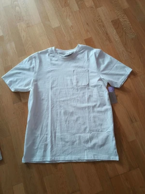 Vit t-shirt med bröstficka - Snygg vit t-shirt från denim day i mjuk bomull. Klassisk passform med rund halsringning och en stilren bröstficka på vänster sida. Perfekt basplagg för en clean och enkel stil.