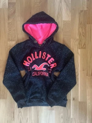 Mörkgrå Hollister hoodie med rosa detaljer - Mysig mörkgrå hoodie från Hollister med stora rosa broderade bokstäver och logga på bröstet. Insidan av huvan är knallrosa och ger en snygg kontrast. Hoodien har känguruficka och dragsko vid huvan. Perfekt för en avslappnad och cool stil.