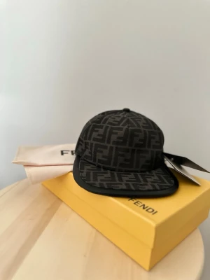 Svart keps med FF-mönster Fendi - Snygg svart keps Fendi med det klassiska FF-logomönstret över hela kepsen. Justerbar baktill med kardborreband och guldfärgad Fendi-detalj. Tillverkad i ett slitstarkt tygmaterial och levereras med originalkartong och dustbag.