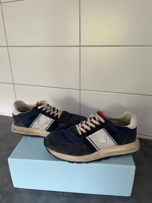 Philippe Model blå sneakers - Snygga sneakers från Philippe Model i marinblått och vitt med grå detaljer. Skorna har en klassisk låg siluett, vita snören och en vit sula. Materialmix av mocka och textil med den ikoniska sköldloggan på sidan. Perfekta för en trendig och avslappnad stil.