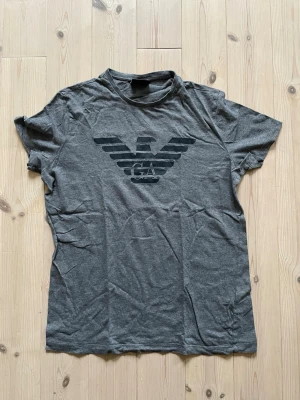Grå t-shirt från Emporio Armani - Snygg grå t-shirt från Emporio Armani med stort svart logotryck på bröstet. Klassisk rund halsringning och korta ärmar. Tillverkad i mjuk bomull. Storlek L men passar mer som S/M. 