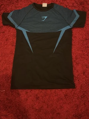 Svart och blå träningströja Gymshark - Snygg svart träningströja från Gymshark med blå detaljer och mönstrad ovandel. Tröjan har korta ärmar och är tillverkad i ett stretchigt funktionsmaterial som andas, perfekt för gymmet eller löpning.