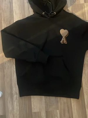 Svart hoodie från Ami Paris med hjärtdetalj - Svart hoodie från Ami Paris med stor huva och klassisk känguruficka. På bröstet sitter ett broderat hjärta med bokstaven A i beige. Hoodien har en avslappnad passform och är tillverkad i mjukt material, perfekt för en chill och stilren look. Har haft på mig den få gånger, Priset kan diskuteras. Ny pris ligger på runt 4200 kr.