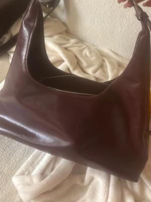 Vinröd axelväska i läder - Snygg vinröd handväska i mjukt läder med enkel och stilren design. Väskan har en bred axelrem och dragkedja upptill. Perfekt för dig som gillar minimalistisk stil och vill ha plats för allt du behöver.